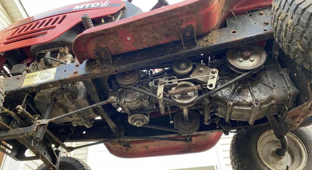 Vue de dessous d'un tracteur tondeuse MTD montrant le système complexe de courroies et de poulies (variateur) qui gère l'avancement avant la boîte de vitesse