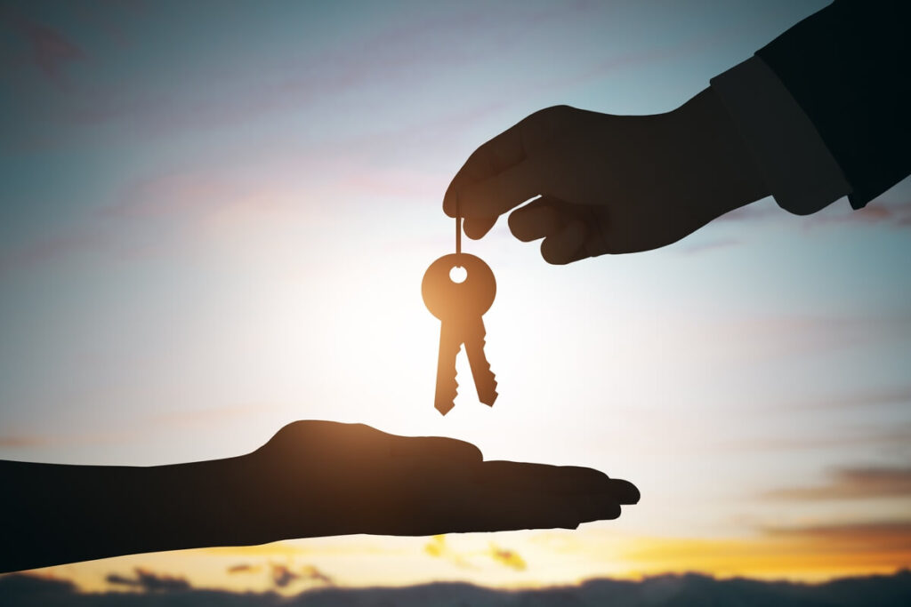 Rêver d'acheter une maison : signification et interprétations 2 Mains tenant un trousseau de clés officiel devant la silhouette floue d'une maison d'habitation.