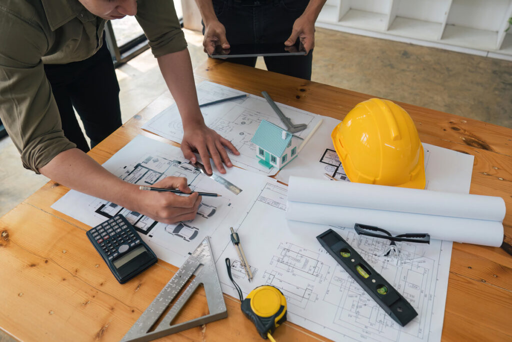 Un maître d'œuvre et ses clients analysant les plans techniques d'une rénovation de maison sur une table de travail.