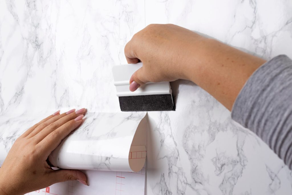 Bricoleur marouflant le premier lé de papier peint intissé avec une spatule plastique pour chasser les bulles d'air.
